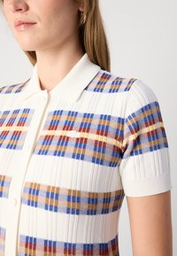 Γυναίκα ντυμένη με ένα άσπρο πλεκτό πόλο πουκάμισο με καρώ σχέδιο σε μπλε, κίτρινο και κόκκινο χρώμα και ριμπ μανίκια κοντά.