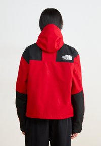 Rote und schwarze Outdoor-Jacke mit Kapuze und strukturiertem Finish; Logo auf dem oberen Rückenbereich; entworfen für Funktionalität und Wärme.
