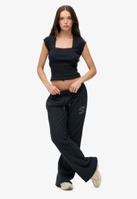 Vrouw die met gekruiste benen staat, gekleed in een zwarte crop top met vierkante halslijn, zwarte Superdry Athletics joggingbroek en beige sneakers, tegen een witte achtergrond.