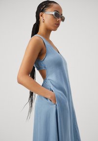 Rochie din denim de culoare albastră deschisă, cu decolteu adânc în V, fără mâneci, cu tăieturi laterale și buzunare. Textură fină și siluetă fluidă.