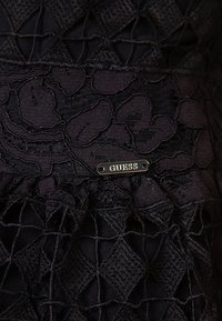 Tessuto nero con sovrapposizione di pizzo floreale e motivo a griglia. Un piccolo tag argento "GUESS" è attaccato in vita.