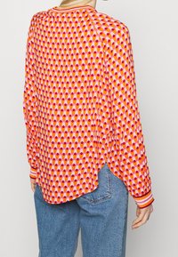 Henkilö, joka on pukeutunut kirkkaan vaaleanpunaisiin, oransseihin ja punaisiin kuvioituihin pitkähihaisiin paitoihin, joissa on raidoitetut hihansuut, yhdistettynä sinisiin farkkuihin, selkä kameraan päin.