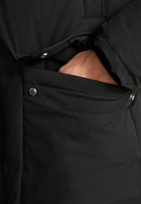 Schwarze isolierte Jacke mit einer seitlichen Tasche und Druckknopfverschluss. Der Stoff ist glatt mit matter Oberfläche und für Wärme entwickelt.