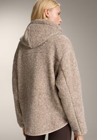 Beżowa, puszysta kurtka z polaru z kapturem, o luźnym kroju i teksturowanej powierzchni. Szwy biegnące wzdłuż ramion i mankietów.