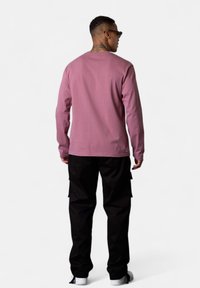 Mann, der sich abwendet, trägt Sonnenbrille, ein langärmliges mauvefarbenes Hemd, schwarze Cargo-Hosen und weiße Sneakers vor einem einfarbigen Hintergrund.