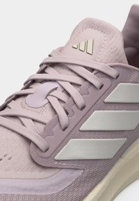 Κοντινό πλάνο ενός ανοιχτού μοβ αθλητικού παπουτσιού με υφασμάτινη υφή, ανοιχτόχρωμα κορδόνια, τρεις λευκές λωρίδες στο πλάι και χρυσό λογότυπο στη γλώσσα.