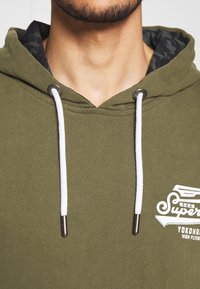 Olivově zelená mikina s kapucí s texturovanou vnitřní podšívkou, bílými šňůrkami a potištěným logem na hrudi. Má kovové detaily.