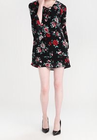 Musta kukallinen mekko, jossa on punaisia ja vihreitä ruusuja, nauhakiinnitys kauluksessa, kolmen neljänneksen hihat ja liehuva helma. Päällä mustat korkokengät.