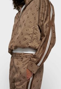 Chándal marrón con un patrón de logotipos repetidos, que incluye una chaqueta con cremallera y pantalones con cintura elástica, con franjas blancas en las mangas y los laterales.