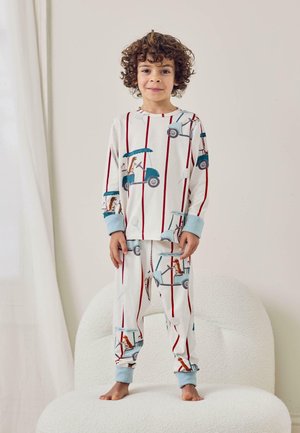 Niño con pelo rizado de pie descalzo sobre una silla blanca texturizada, vestido con pijama con carros de golf y perros marrones sobre un fondo blanco con rayas rojas.