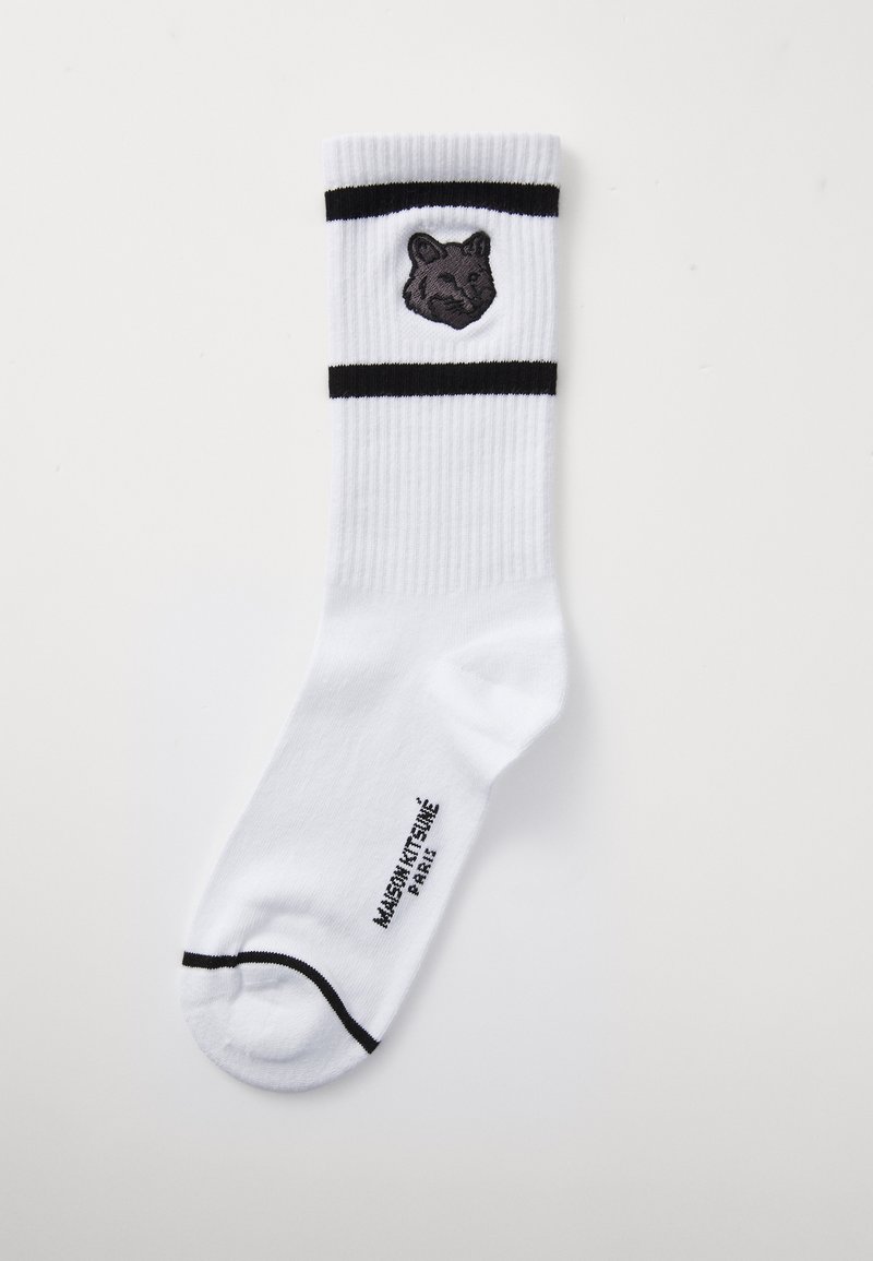 Chaussettes blanches en coton avec une broderie de tête de lion grise, bordées de rayures horizontales noires près du haut et avec le nom de la marque imprimé sur la semelle.