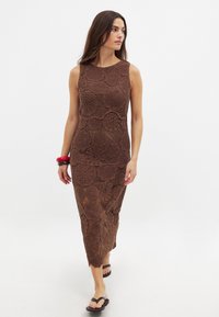 Abito lungo all'uncinetto marrone con motivo floreale, senza maniche e con uno spacco laterale. Modello abbinato a flip-flop nere e bracciali rossi.
