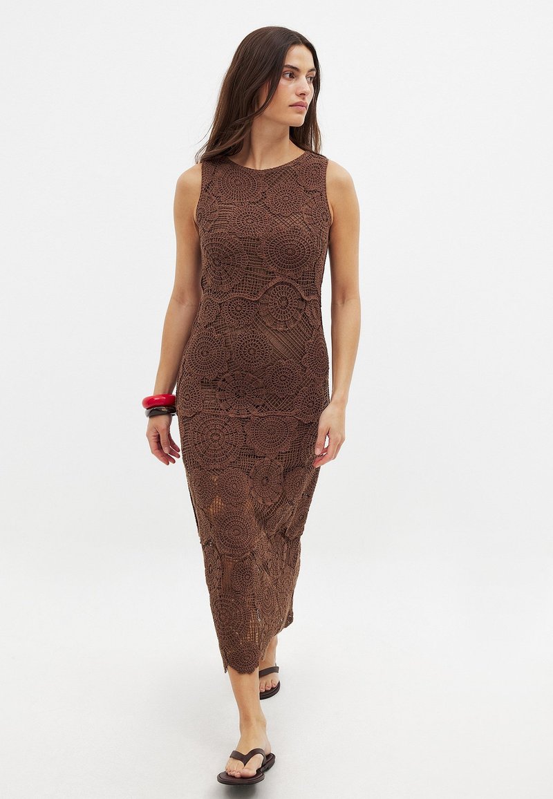 Abito lungo all'uncinetto marrone con motivo floreale, senza maniche e con uno spacco laterale. Modello abbinato a flip-flop nere e bracciali rossi.