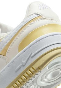 Zapatilla blanca y dorada con un upper de cuero suave, suela de goma, acentos texturizados y un logotipo elevado en el talón.