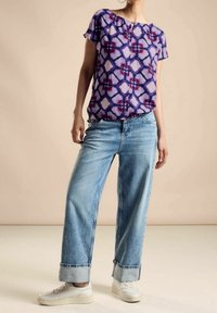 Blusa viola a fantasia con scollatura a chiave, abbinata a jeans blu chiaro a gamba larga e sneakers bianche. Tessuto morbido.