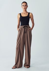 Pantaloni ampi marroni con vita elasticizzata e dettaglio con cordoncino, abbinati a un top aderente nero. Design minimalista e texture liscia.