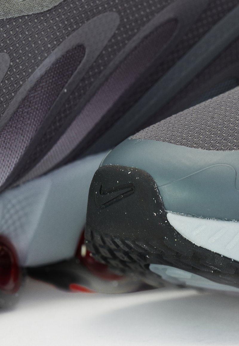 Zapatillas deportivas grises con parte superior de malla texturizada, talón curvado, suela de goma negra y acentos rojos en la suela, con un logo en el talón.