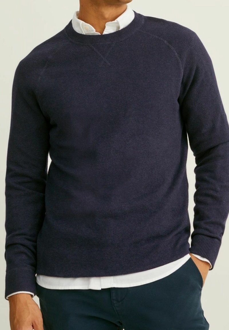 Marinblå stickad sweatshirt med ett texturerat mönster, rundad halsringning och raglanärmar. Bärs över en vit skjorta med krage.