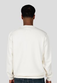 Felpa bianca con una texture liscia, polsini a coste e scollatura rotonda. Presenta una cucitura centrale e un piccolo etichetta sul retro.