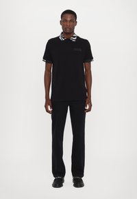 Black polo shirt with striped collar, short sleeves, and logo detail. Paired with black trousers and shoes. Simple, casual design.