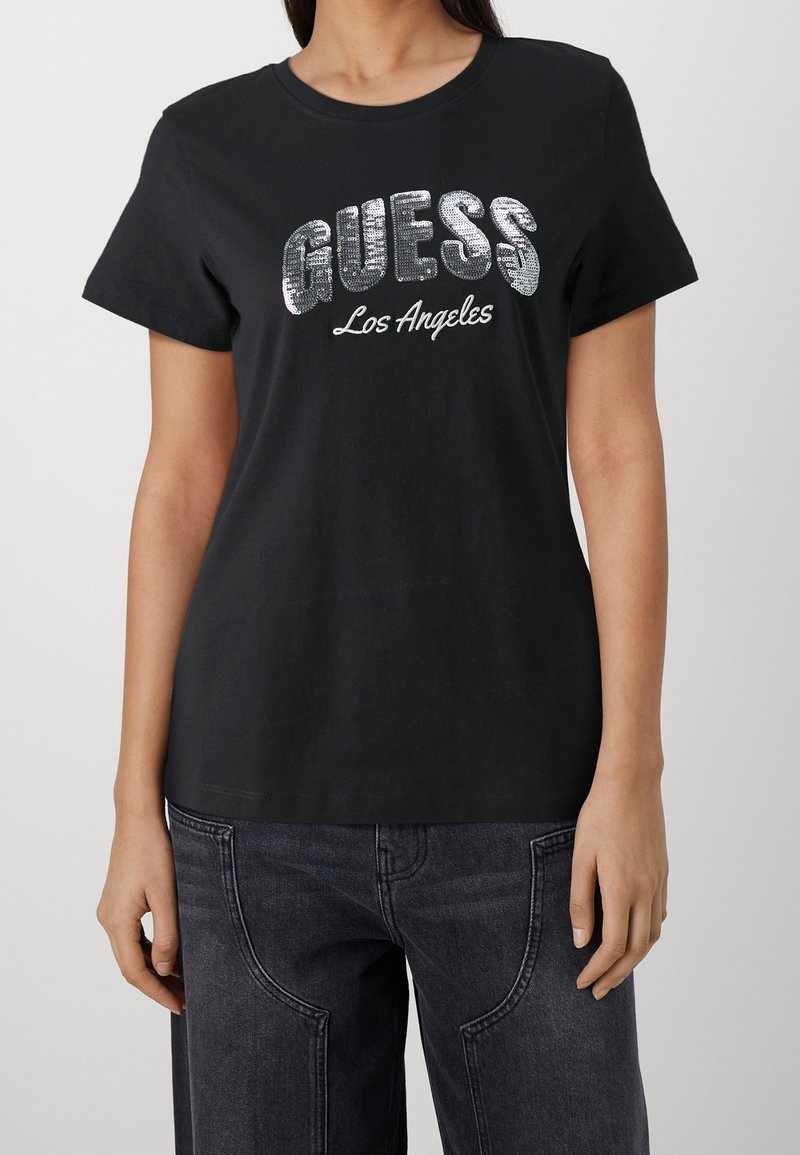 Sort bomulds T-shirt med sølvfarvede pailetter, der danner "GUESS" logoet med teksten "Los Angeles" nedenfor. Korte ærmer og rund halsudskæring.