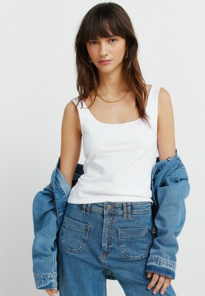 Młoda kobieta o długich ciemnych włosach, nosząca złote kolczyki koła, złoty naszyjnik na łańcuszku, biały bezrękawnik, jeansy i kurtkę jeansową zsuniętą z ramion.