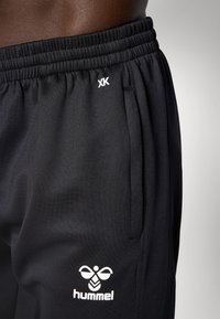 Közelkép egy személyről, aki fekete Hummel márkájú sportnadrágot visel elasztikus derékkal és oldalzsebbel, amin a márka logói láthatók.