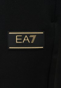 Μαύρο υφασμάτινο με ανάγλυφη υφή, με ορθογώνιο μαύρο και χρυσό πατς που εμφανίζει το λογότυπο "EA7 Emporio Armani".