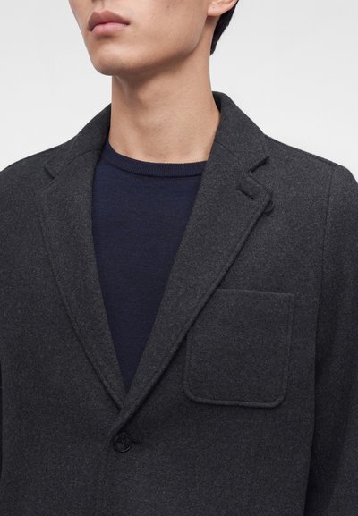Homme portant un blazer gris foncé sur un pull bleu marine à col rond, montrant le revers du blazer, la poche poitrine et un seul bouton.