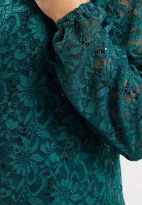 Błękitna koronkowa bluzka z kwiecistymi wzorami, cekinami i elastycznymi mankietami. Przezroczysta tekstura podkreśla szczegóły projektu i głębię.