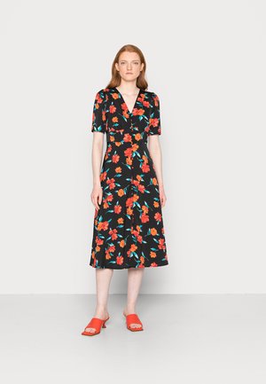 Vestido preto floral com mangas curtas, apresentando flores vermelhas e turquesas. Modelo a usar sandálias deslizantes vermelhas. O vestido tem um decote em V e um corte até meio da coxa.