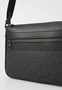 Black shoulder bag with textured fabric, smooth leather accents, silver zipper, and embossed logo detail on the front. Adjustable strap included.
