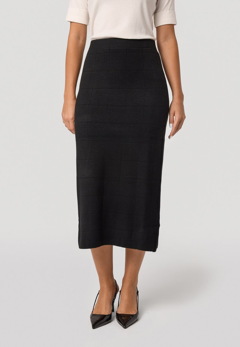 Jupe crayon noire avec un motif à carreaux texturé, longueur mi-mollet, taille haute, associée à des talons aiguilles noirs.