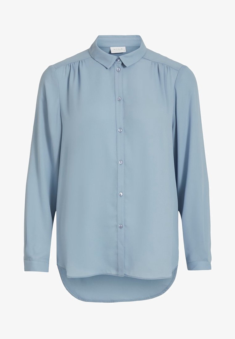 Blouse bleu clair à manches longues avec patte de boutonnage à l'avant, col classique, légers fronces aux épaules, et ourlet arrondi asymétrique.