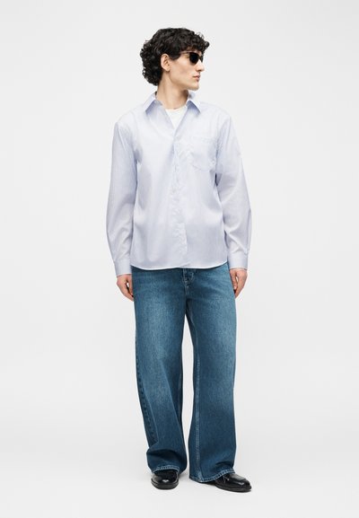 Νέος άνδρας με σγουρά μαλλιά φορώντας γυαλιά ηλίου, ένα ανοιχτό μπλε πουκάμισο με ρίγες, φαρδιά μπλε τζιν και μαύρα παπούτσια, στέκεται κοιτάζοντας πλάγια.