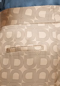 Smėlio spalvos kelnės, pagal pasikartojantį kontūrinių figūrų raštą. Juosmuo dekoruotas mažomis kišenėlėmis gale ir klostėmis.