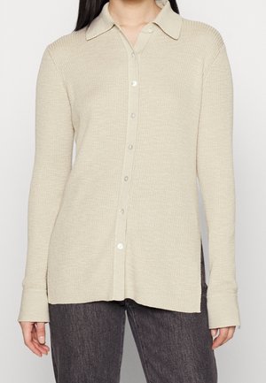 Femme portant une chemise beige côtelée à manches longues boutonnée avec col et un pantalon gris foncé, debout, bras détendus sur les côtés.