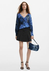 Top lungo blu a maniche lunghe con motivo a fantasia e scollatura a V, abbinato a una gonna nera plissettata e a una borsa in denim. Tacchi neri con cinturini completano il look.