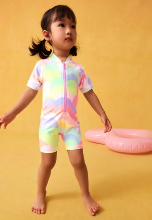 Nuori lapsi, jolla on lettikiharat, pukeutuneena pastellinväriseen sateenkaarenkuvioiseen lyhythihaiseen uimapukuun. Lapsi seisoo paljain jaloin keltaisella lattialla kahden vaaleanpunaisen ilmatäytteisen renkaan vieressä.