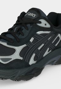 Μαύρο και γκρι αθλητικό παπούτσι Asics με υφασμάτινο δίχτυ, συνθετικές επικαλύψεις και κλείσιμο με κορδόνια, εμφανιζόμενο από κοντινή γωνία εμπρός-πλευράς.