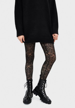 Kvinde iført sorte strømpebukser med blondermønster, sort oversized sweater og sorte snørestøvler, stående mod en ensfarvet baggrund.