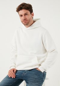 Hvid hættetrøje med kængurulomme, lavet af blødt stof. Har ribkantede manchetter og en afslappet pasform. Kombineret med blå jeans.