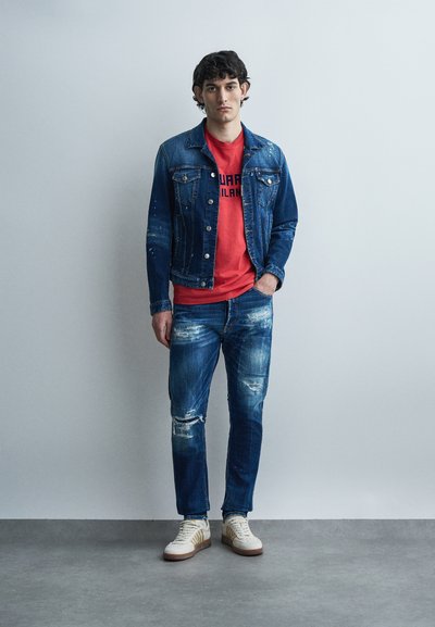 Jeune homme debout contre un mur uni, portant une veste en jean usée, un t-shirt rouge à motif, un jean déchiré, et des baskets blanches.