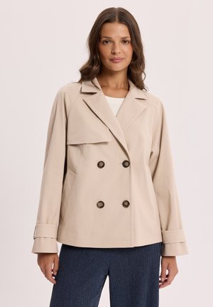 Kobieta ubrana w beżową dwurzędową marynarkę z dużymi guzikami na białej bluzce i ciemnych teksturowanych spodniach, stojąca na jednolitym tle.