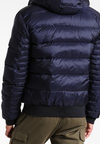 Granatowa puchowa kurtka z kapturem, z pikowanymi sekcjami i ściągaczami przy mankietach. Gładki, błyszczący materiał o praktycznym kroju.