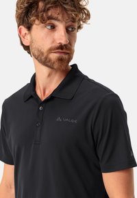 Černé polo tričko s klasickým límečkem, obsahující zapínání na knoflíky a decentní logo Vaude na hrudi. Vyrobeno z hladké, prodyšné tkaniny.