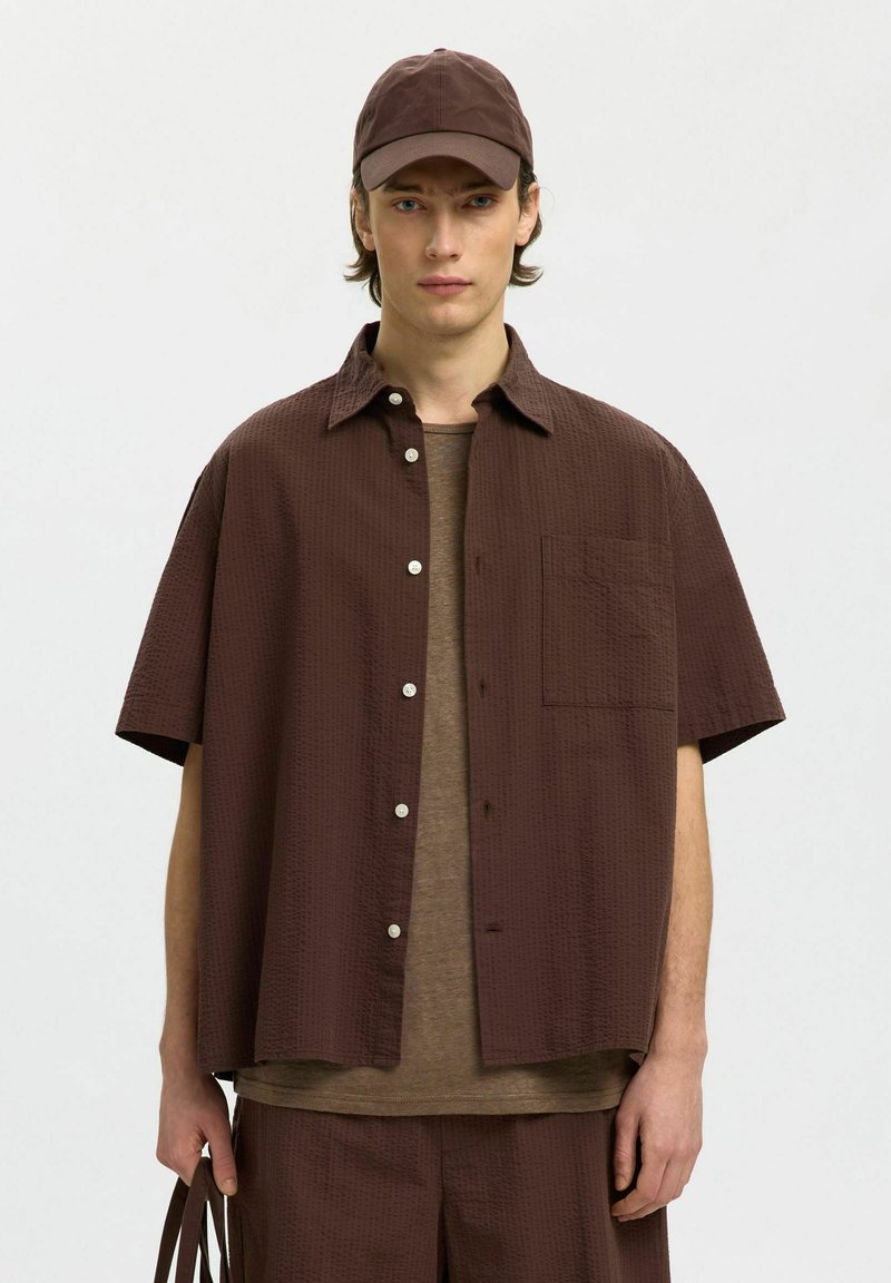 Jeune homme portant une casquette marron foncé, une chemise à manches courtes boutonnée, un short assorti, tenant un sac marron sur un fond uni.