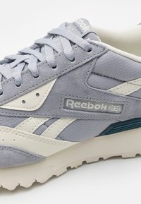 Šedá semišová teniska s bílými doplňky, texturovaným svrškem a gumovou podrážkou. Obsahuje logo Reebok a tradiční zavazování na tkaničky.