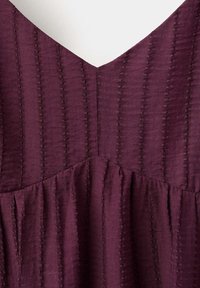 Gros plan d'un tissu violet texturé avec des lignes de couture verticales formant un col en V et un détail de couture froncée sous la poitrine.