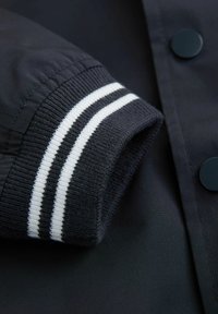 Puño de chaqueta negra con rayas blancas, con un tejido texturizado. El material es suave, con un detalle de botón oscuro visible cerca.